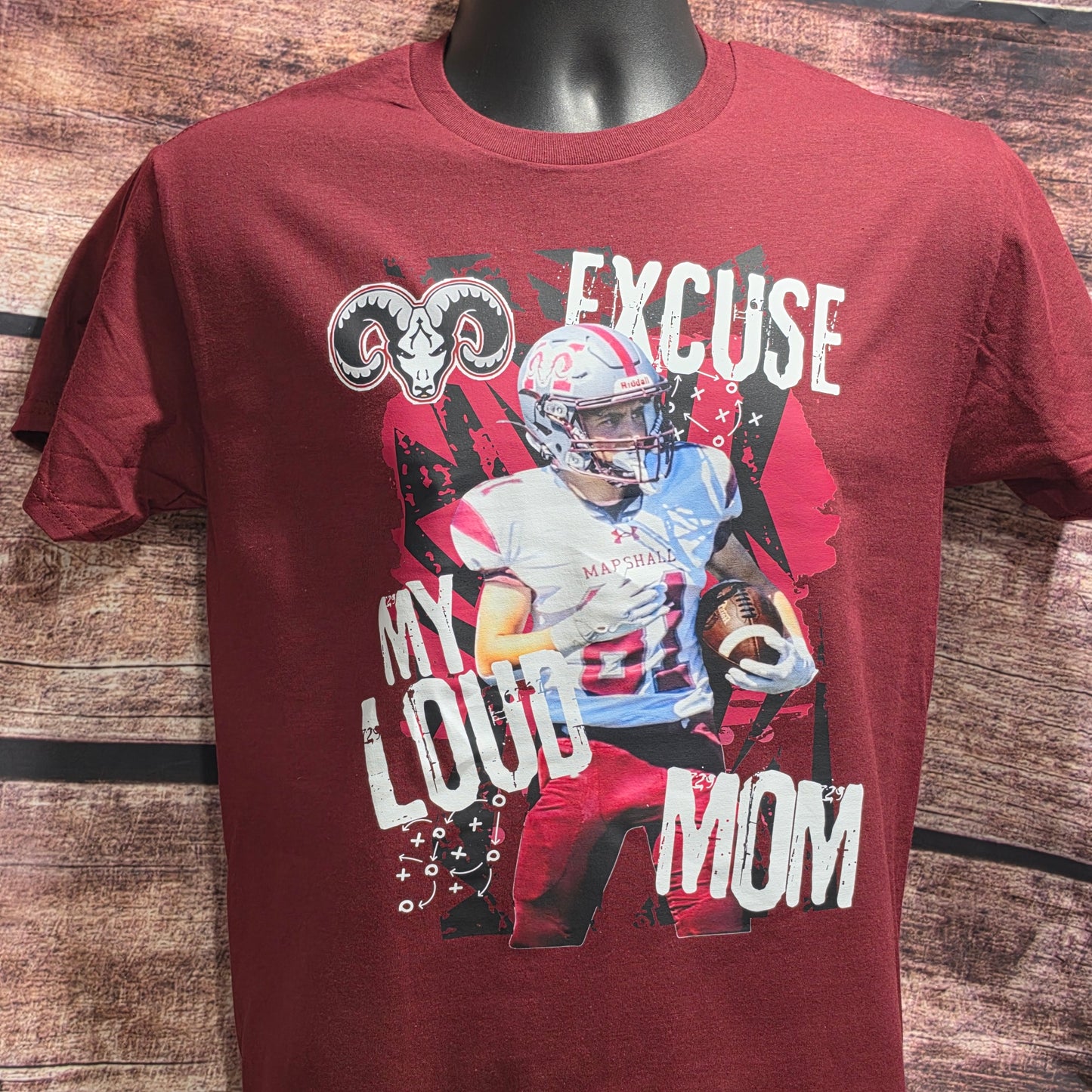 A maroon short-sleeve graphic t-shirt displayed on a black mannequin torso against a rustic wood plank background. The graphic features a football player, a ram head logo, and the text 'MY LOUD MOM' and 'EXCUSE MY LOUD MOM' in distressed white lettering.