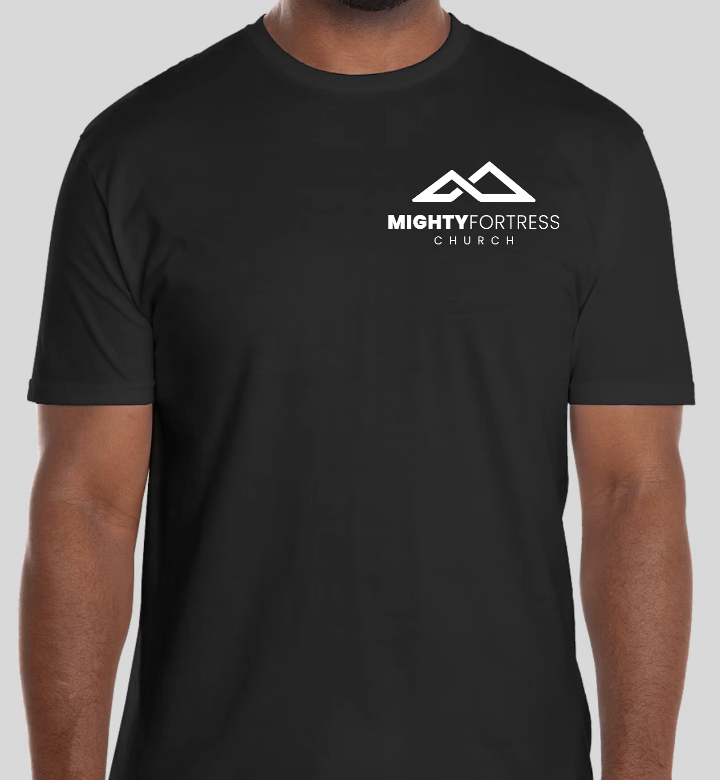 A man wearing a black short-sleeve crewneck t-shirt with a white 'MIGHTY FORTRESS CHURCH' logo printed on the left chest, shown against a solid gray background.