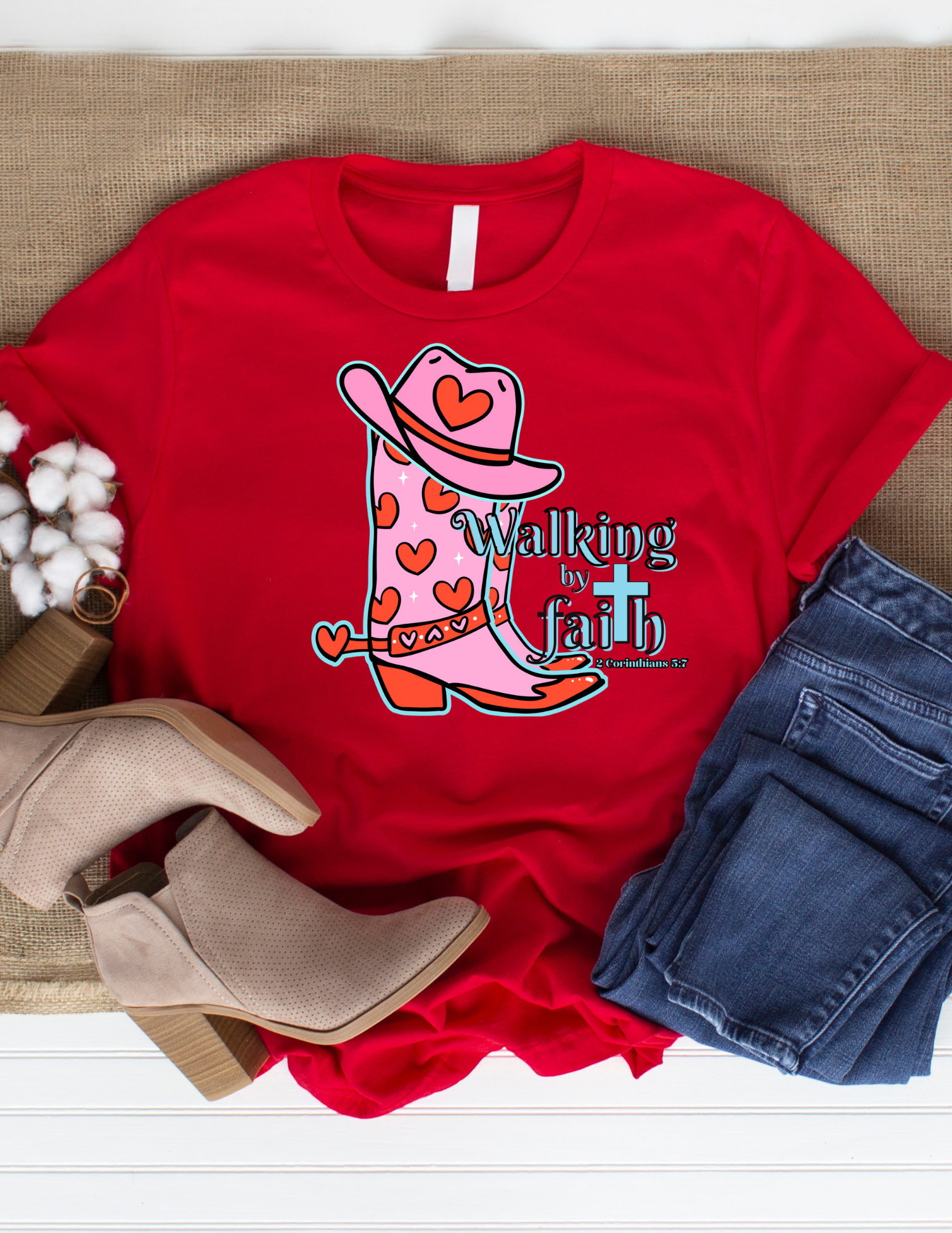 Red t-shirt with pink cowboy boot graphic and 'Walking by Faith' text, displayed with boots and jeans on a wooden surface.
