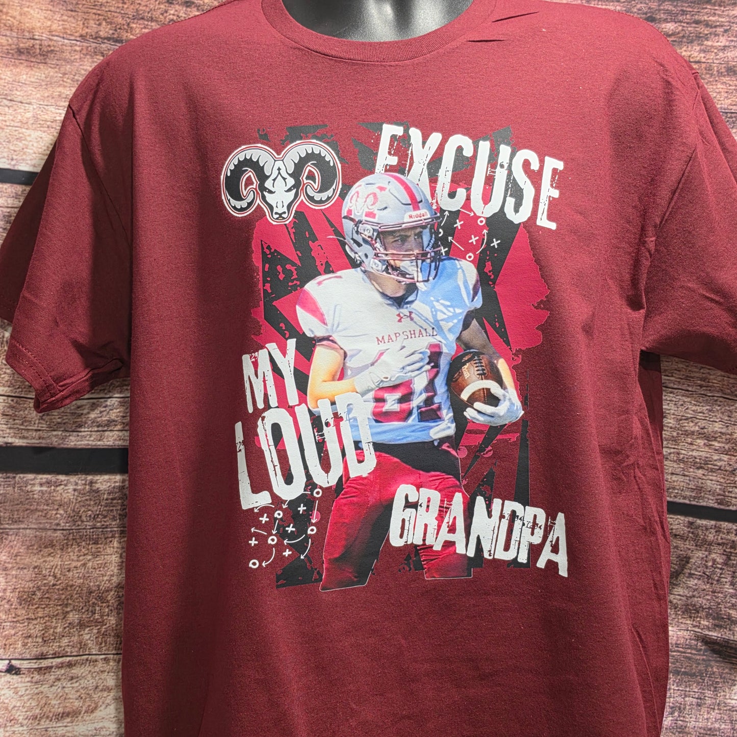 A maroon short-sleeve graphic t-shirt displayed on a mannequin torso against a rustic wooden plank background. The graphic features a football player in a white and red uniform with the text 'MY LOUD GRANDPA' and a ram skull logo.