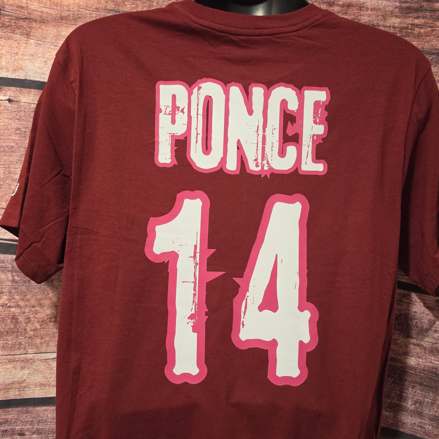 A dark red short-sleeve t-shirt displayed on a black mannequin torso against a rustic wooden plank background. The back of the shirt features the word 'PONCE' and the number '14' printed in distressed white with a pink outline.